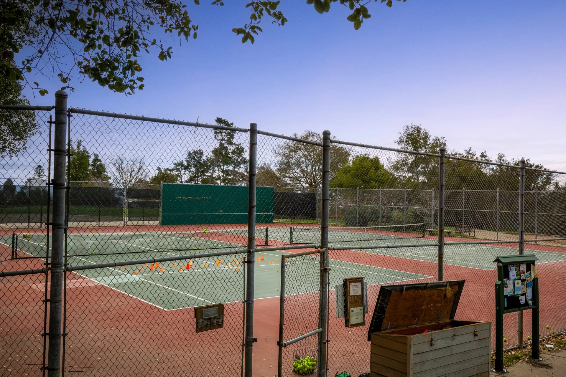 An empty outdoor tennis court surrounded by a chain-link fence with trees and a blue sky in the background. Tennis balls, cones, and informational signs are visible near the fence.