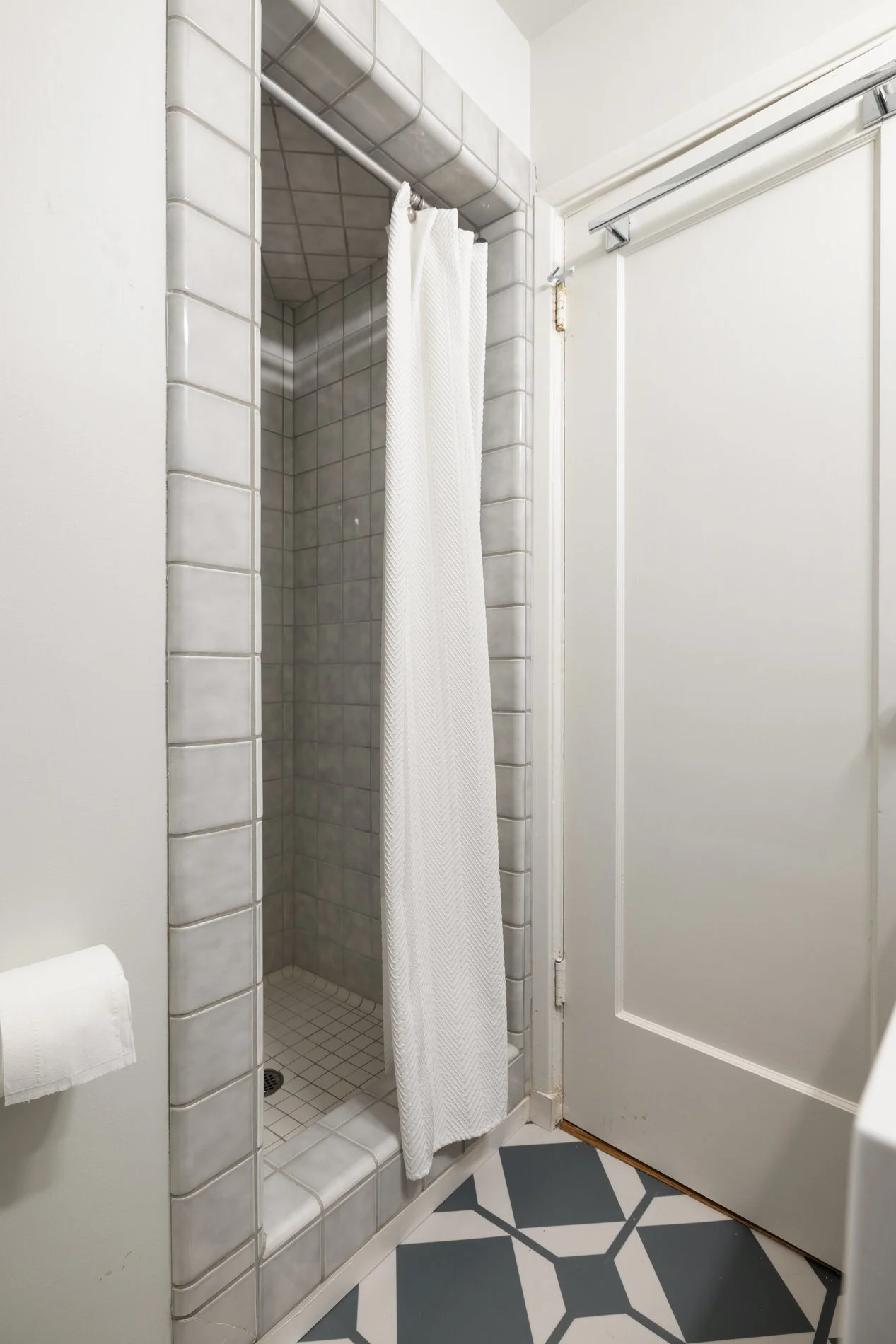 Small shower with gray tiled interior, white curtain, white towels, and a sliding door, next to a white wall with patterned tile floor.