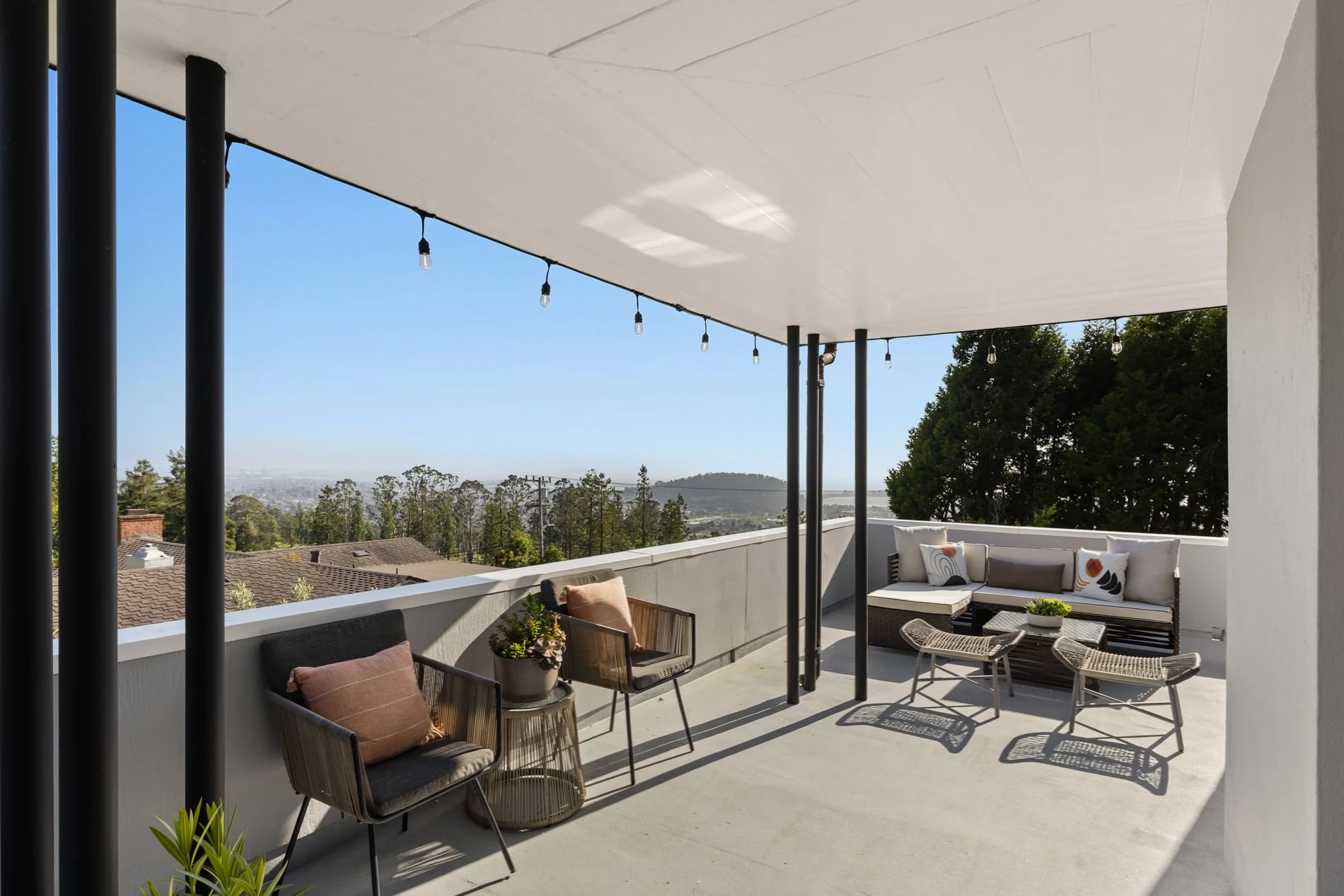 Modern rooftop patio with seating area: black chairs with pillows, wicker chairs, small plants, and string lights, overlooking trees, rooftops, and distant hills under a clear sky.