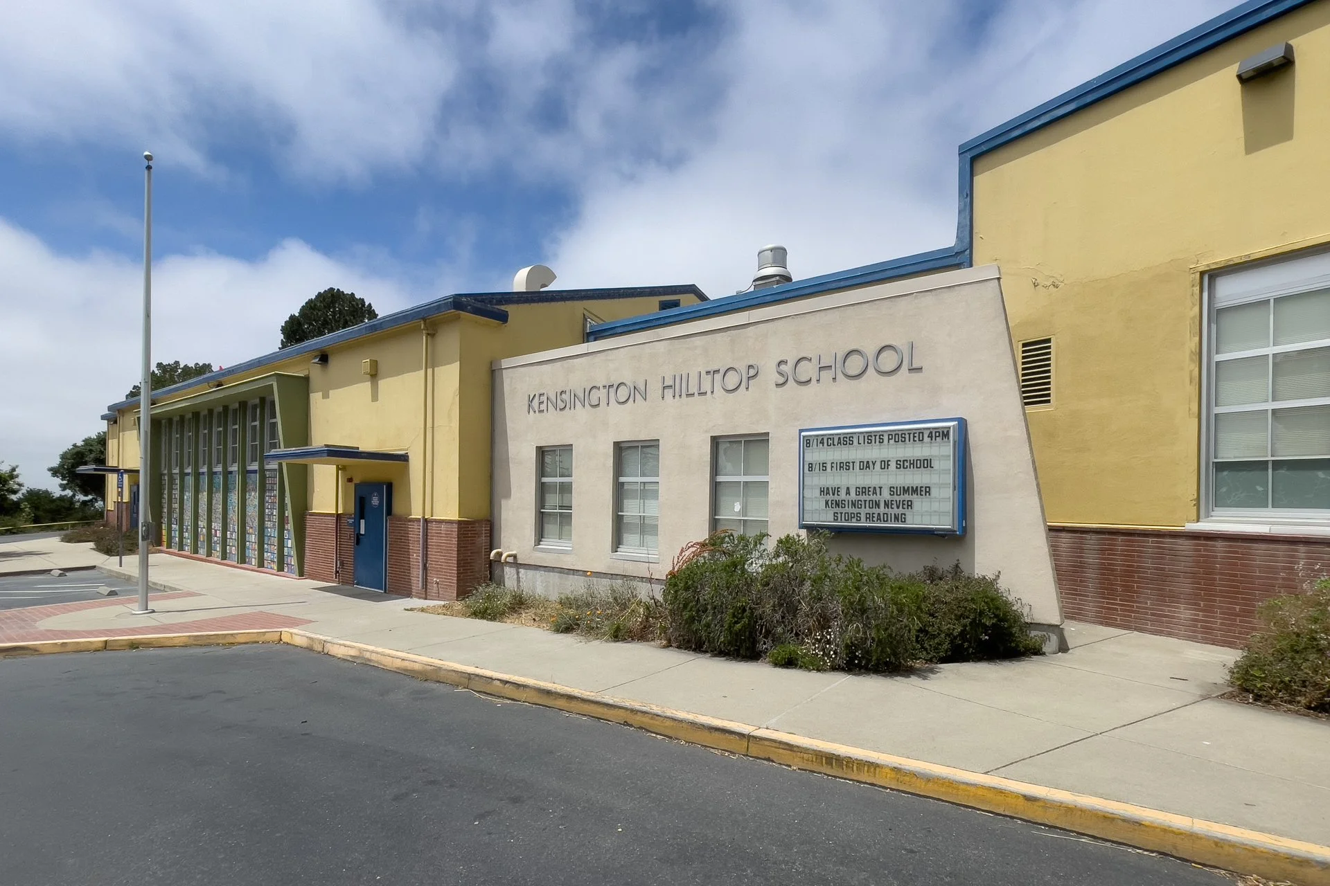 Front view of Kensington Hilltop School building with a signboard, a flagpole, and a parking lot in the foreground, under a partly cloudy sky.