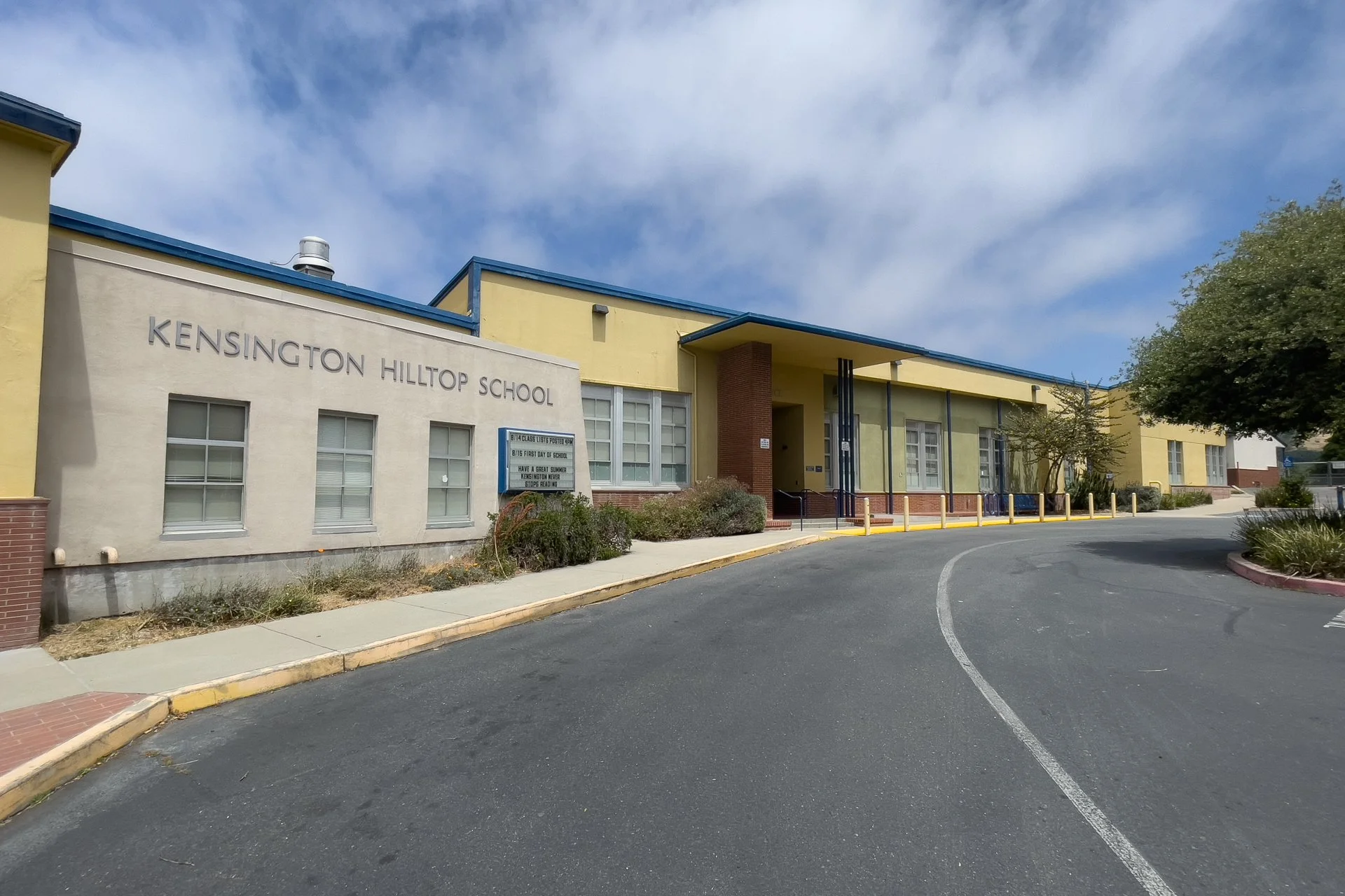 Exterior of Kensington Hilltop School building with a yellow and beige facade, a blue roof, a signboard, and trees nearby, under a partly cloudy sky.