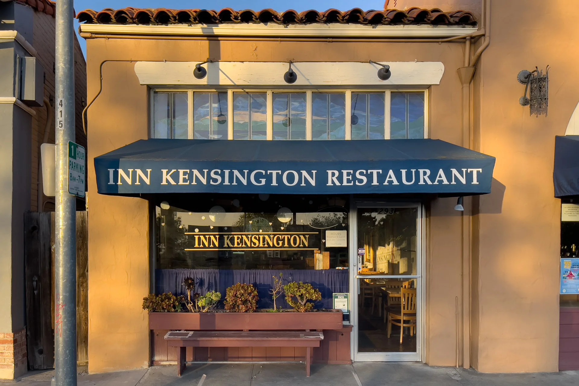 The storefront of a restaurant called 'Inn Kensington' with a blue awning and large windows. There is a plant box with flowers outside and a bench. A sign on the door displays the restaurant hours and a laminated card on the door shows 'Pass'.