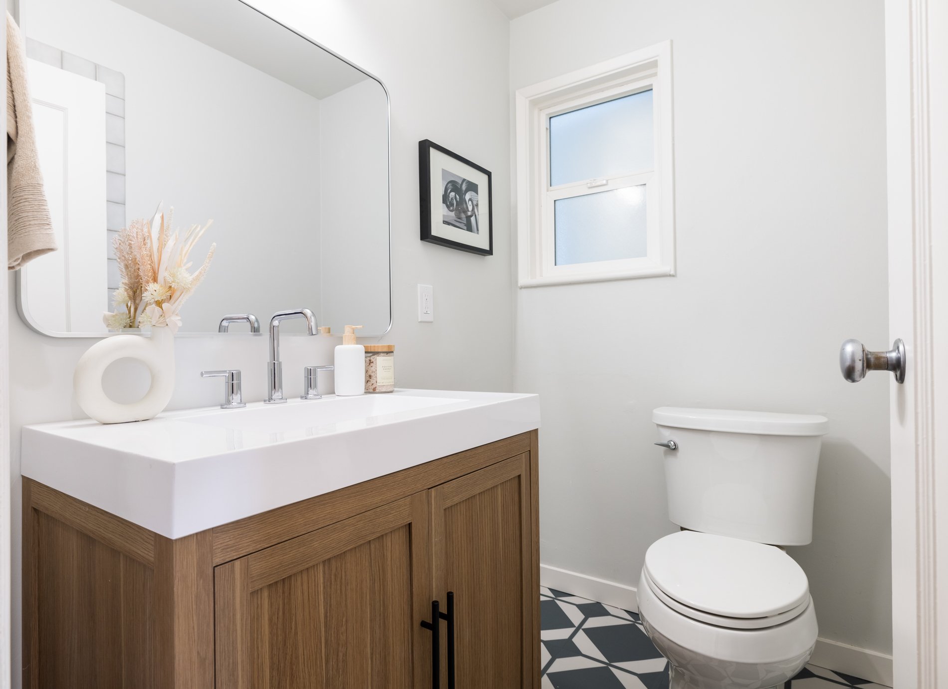 A modern bathroom featuring a wooden vanity with a white countertop, a rectangular mirror, neutral-colored decor including a vase with dried flowers, and black and white geometric patterned floor tiles. There is a window allowing natural light and a 