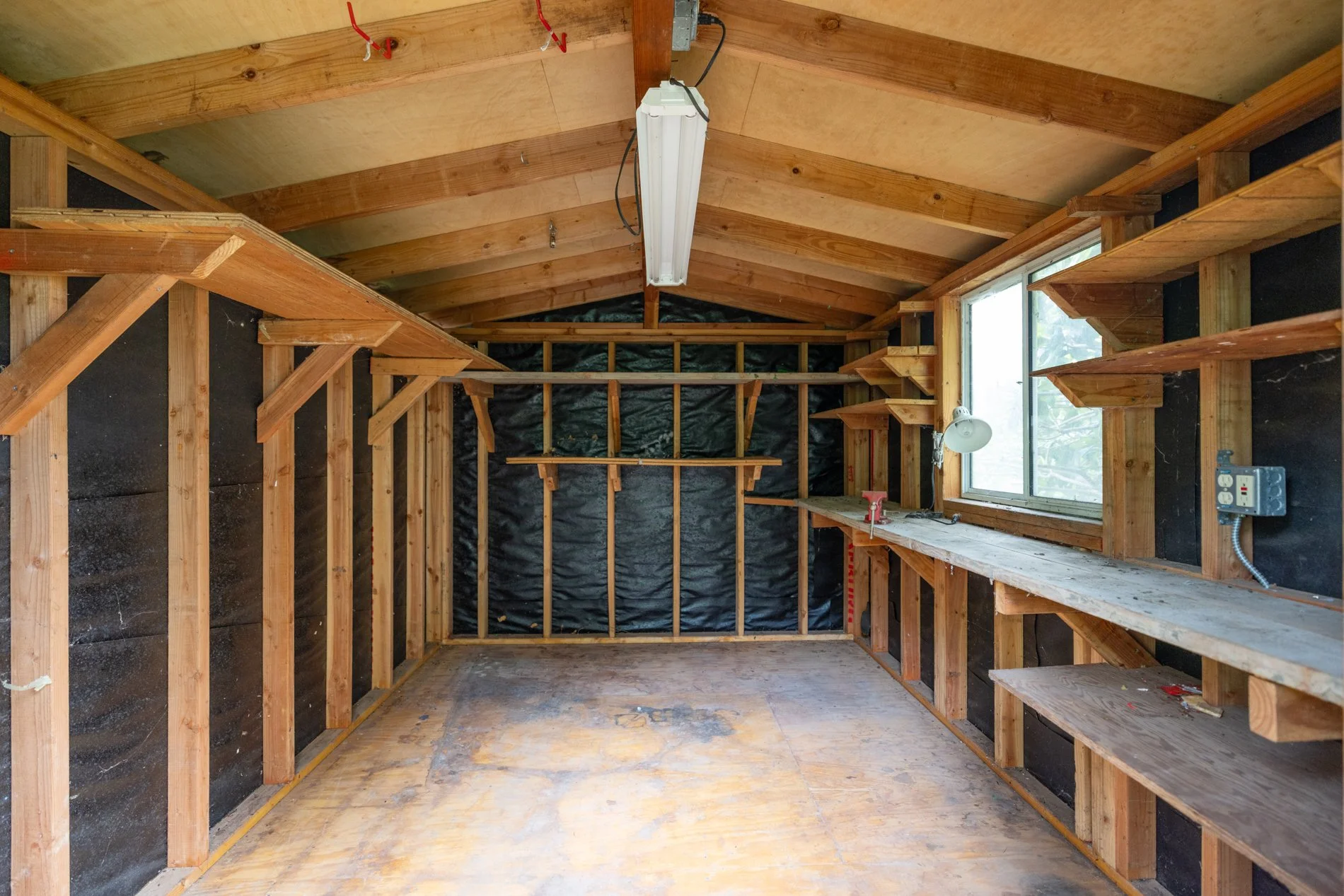Inside a partially constructed room with exposed wooden framing, a workbench, shelves, a window, and a fluorescent light fixture on the ceiling.