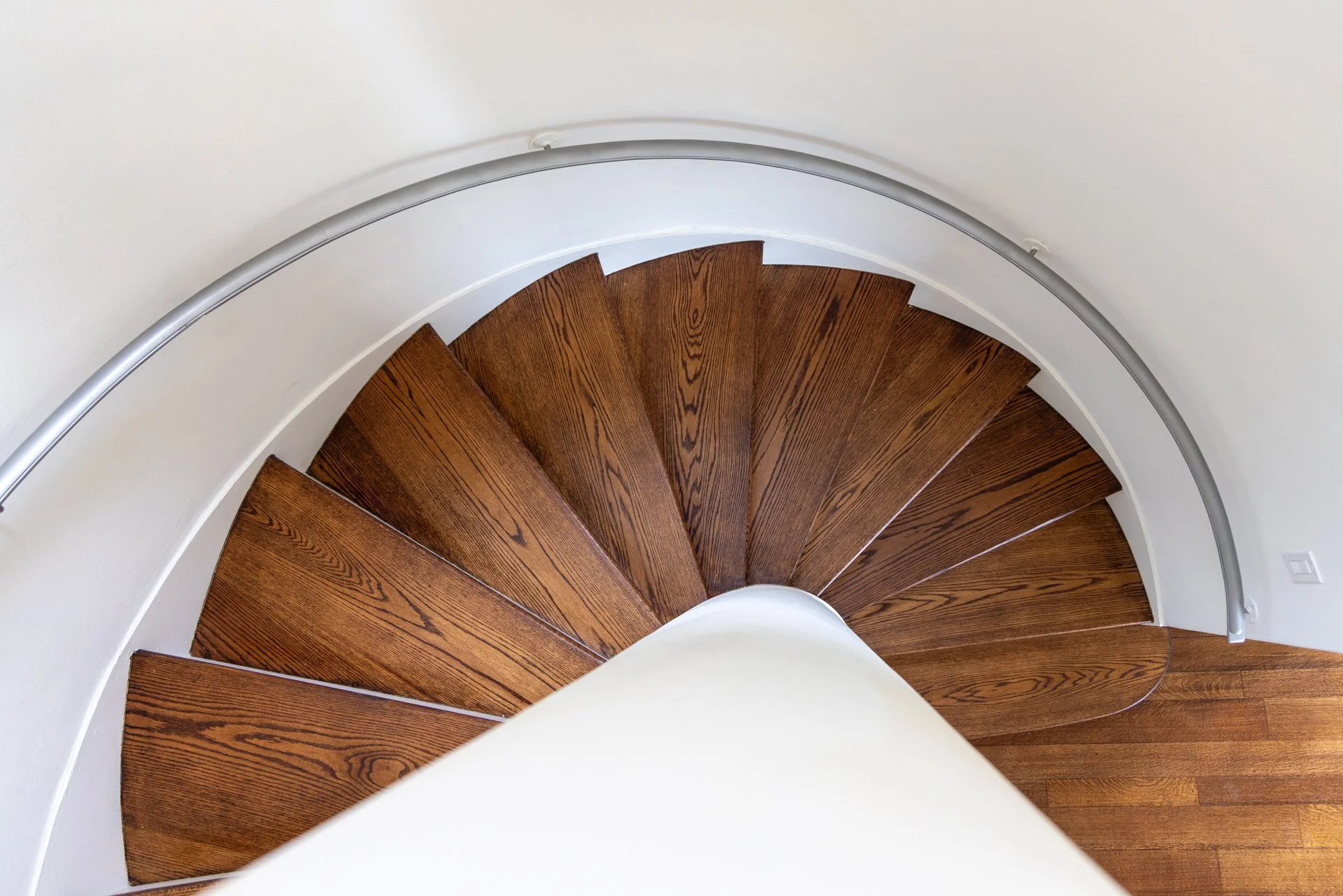 Top-down view of curved wooden staircase with metal railing and white walls.