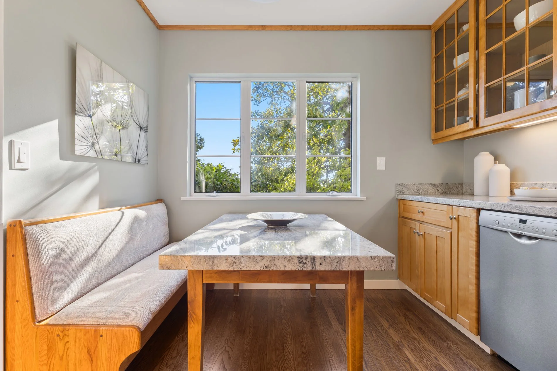 A kitchen dining area with a marble-top table and a bench with beige upholstery along the wall. There is a large window showing green trees outside, with sunlight streaming in. Wooden cabinets with glass doors and white dishware are on one side, whil