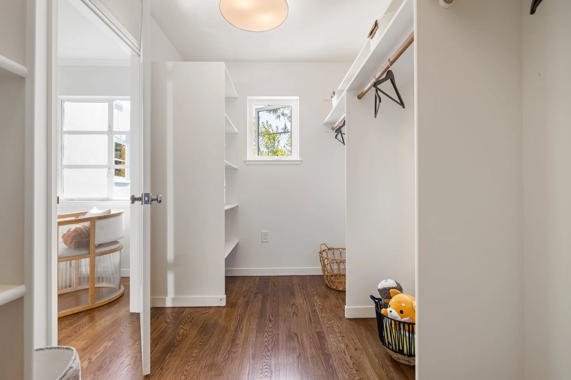 Empty walk-in closet with white walls, hardwood floor, built-in shelves, and a small window showing trees outside. There are some plush toys in a basket and a laundry basket with a stuffed animal.