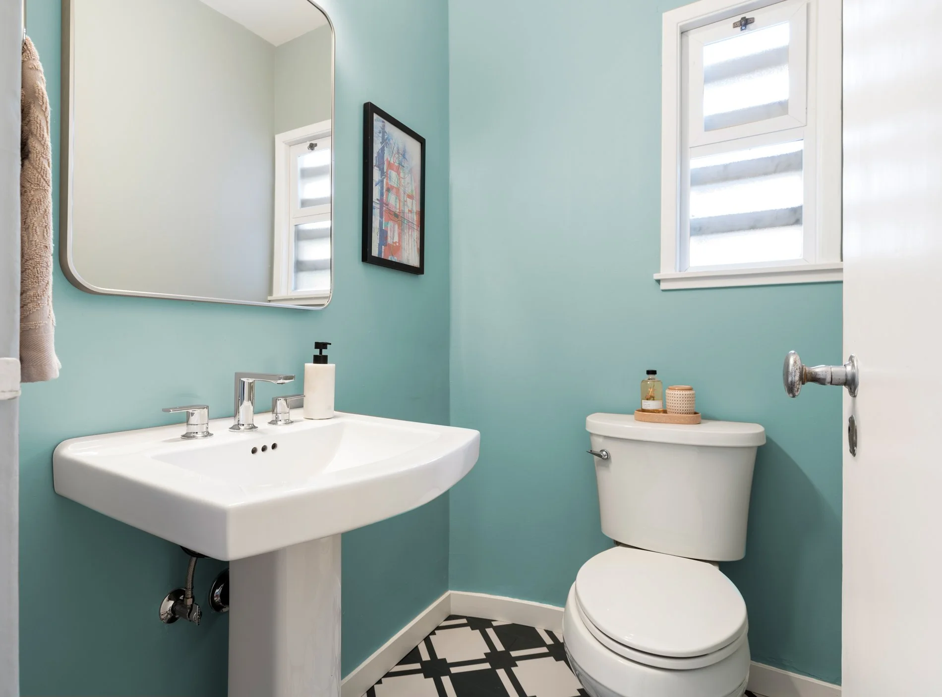 Bathroom with a light blue wall, white sink with chrome fixtures, mirror, framed artwork, window with horizontal blinds, toilet with decorative items on tank, patterned floor tiles, and towel hanging on wall.
