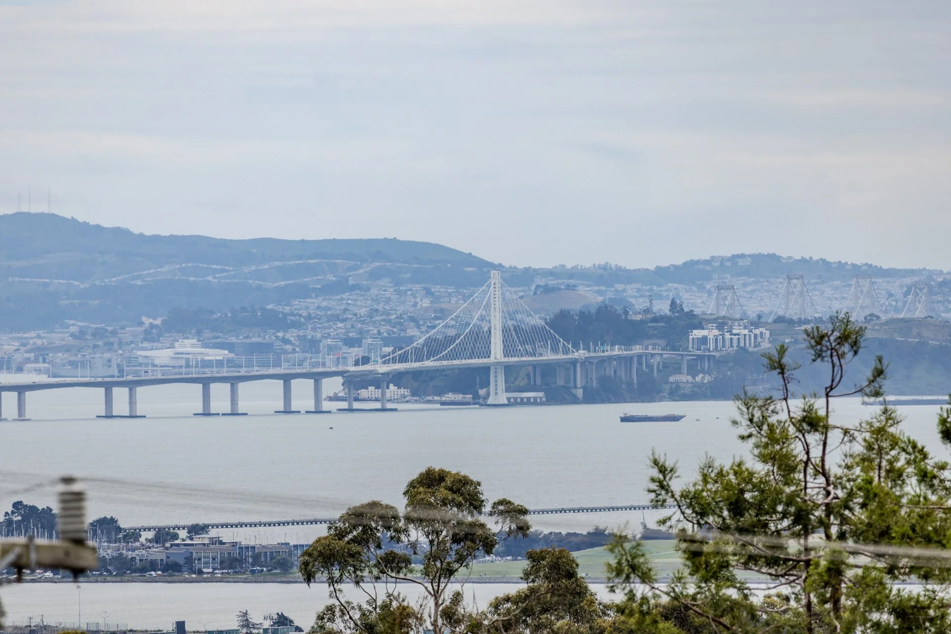A large body of water with a bridge spanning across it, mountains and hills in the background, and trees in the foreground.