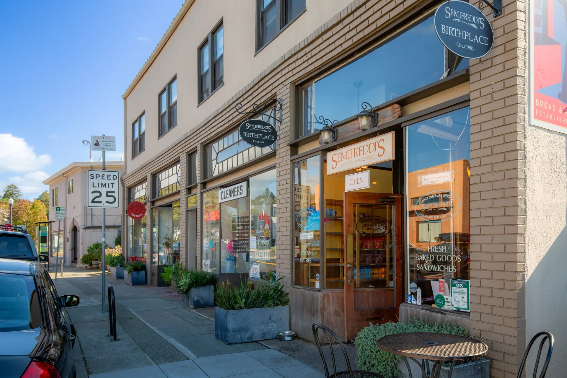 Exterior view of a bakery and shop called Semifreddi's Birthplace with large windows displaying signs for baked goods and sandwiches. Potted plants line the sidewalk, and a few parked cars are visible, along with a speed limit sign of 25 miles per ho