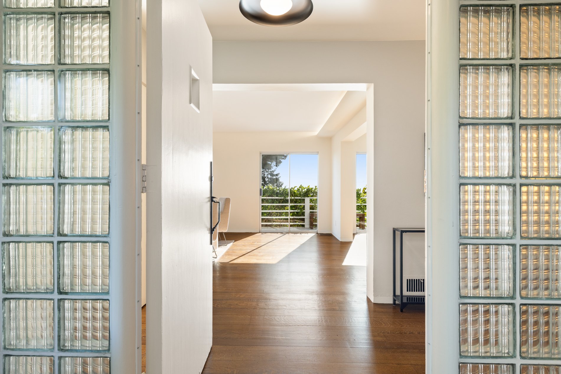 Interior view of a modern apartment with wooden flooring, white walls, large windows, and a balcony with a view of greenery outside. Glass block walls on each side frame the entrance.