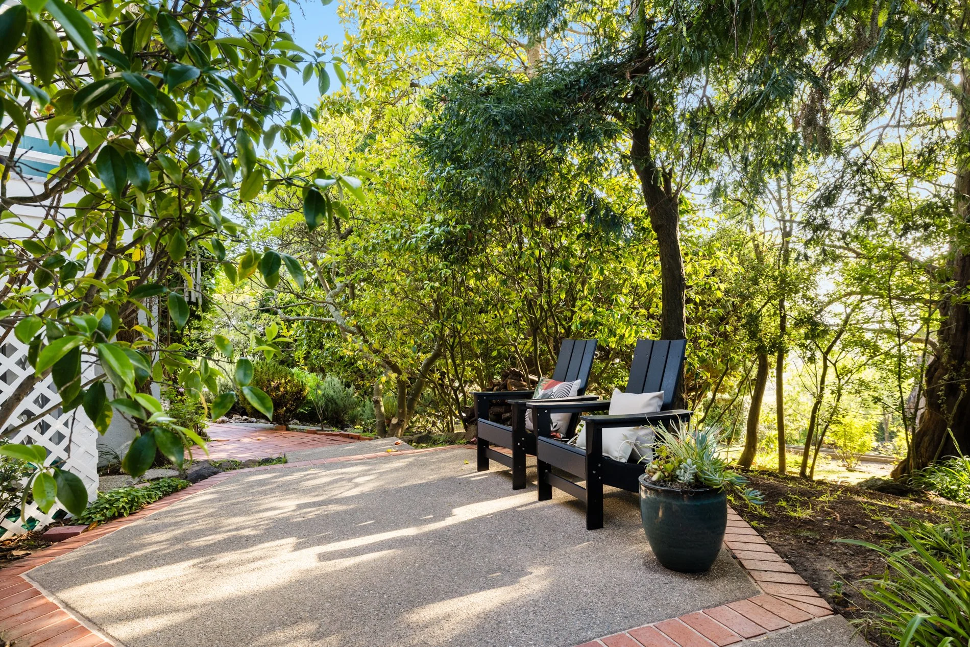 A backyard patio with two black chairs, a potted plant, and lush green trees providing shade.