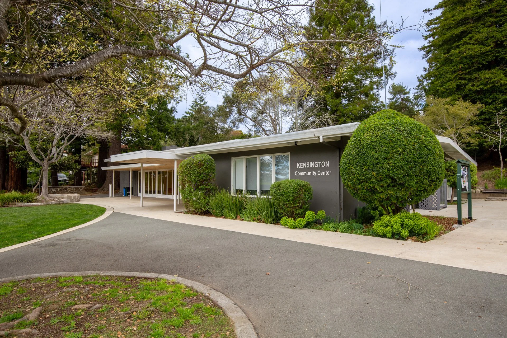 The Kensington Community Center is a single-story building with a gray exterior, large front windows, and a white flat roof. There is a curved asphalt walkway leading up to the entrance, surrounded by well-maintained grass and lush green plants. Two 