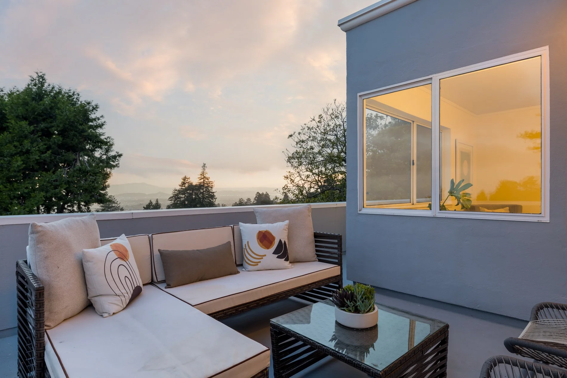 A balcony outdoor seating area with a cushioned sofa and pillows, overlooking trees and a sunset sky, with a large window showing a cozy interior illuminated with warm light.