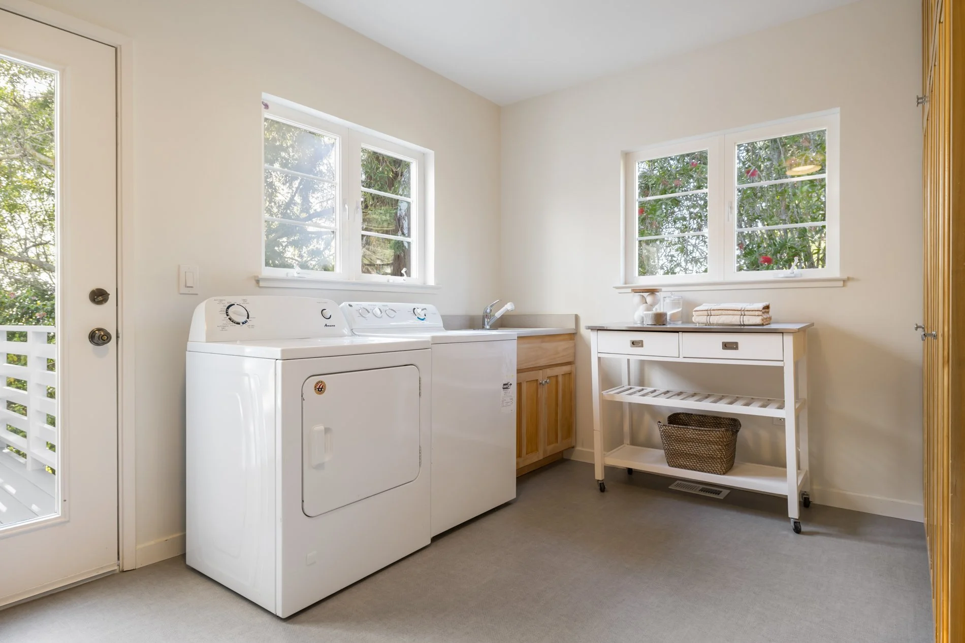 Laundry room with white washing machine and natural wood cabinets, white table with folded towels, potted plants, and windows showing outside greenery.