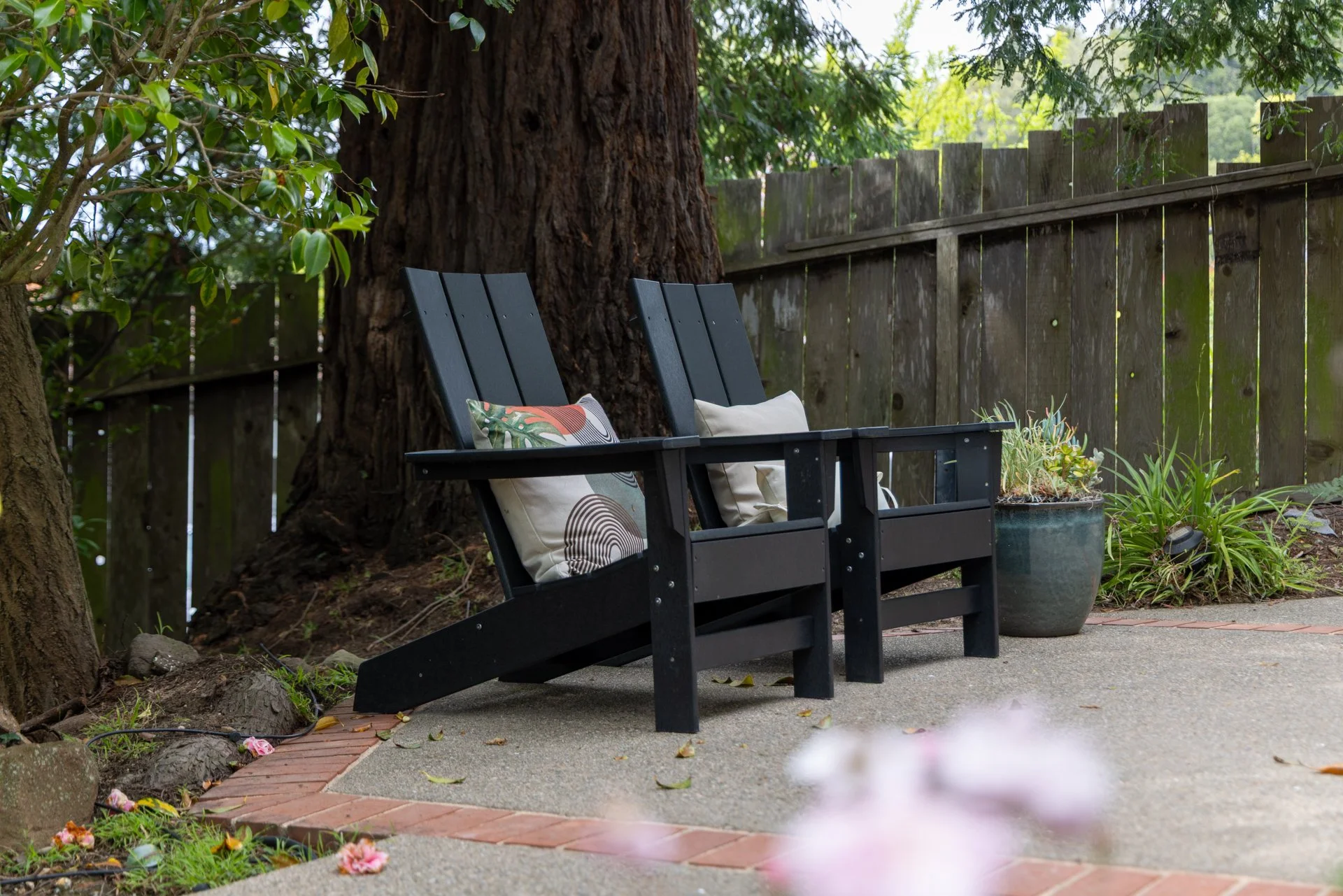 Black wooden bench with two beige cushions, situated in a garden with a large tree, wooden fence, potted plants, and brick-edged pathway, during daytime.