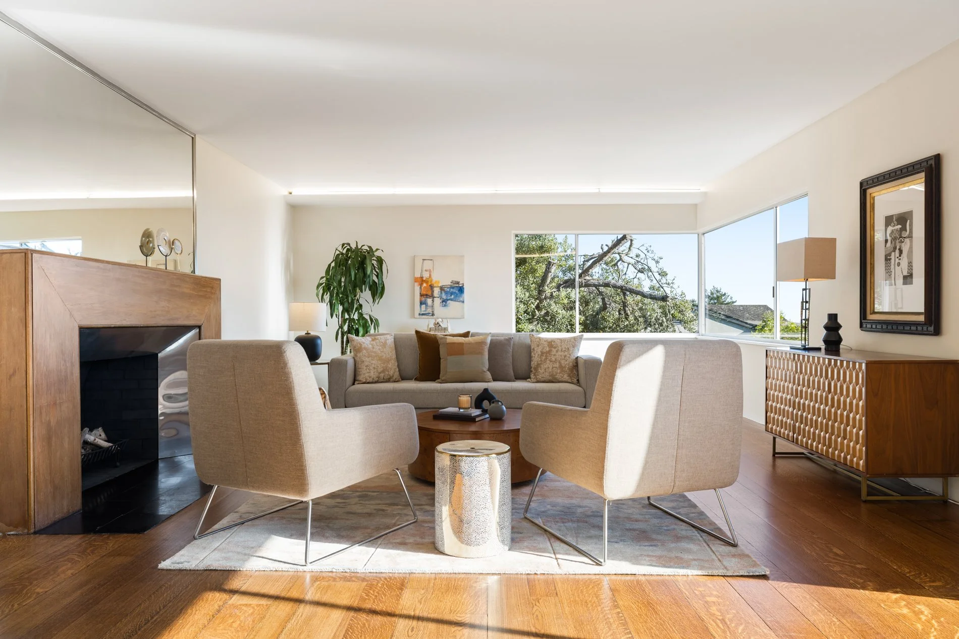 Living room with large window view, beige sofa, armchairs, wooden coffee table, artwork on walls, and hardwood floors.