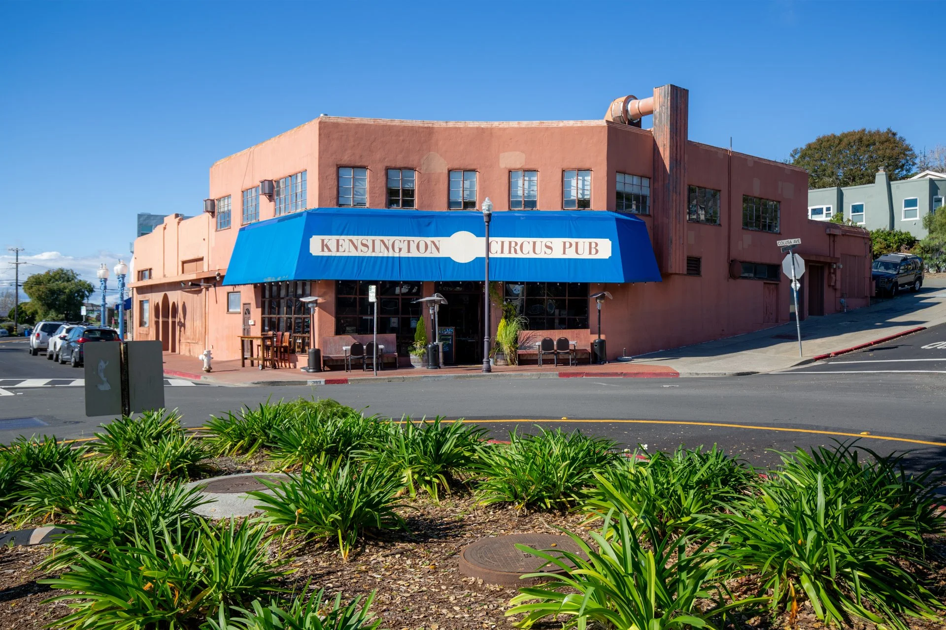 Building with a blue awning that reads 'Kensington Circus Pub', with outdoor seating and street signs at the corner.