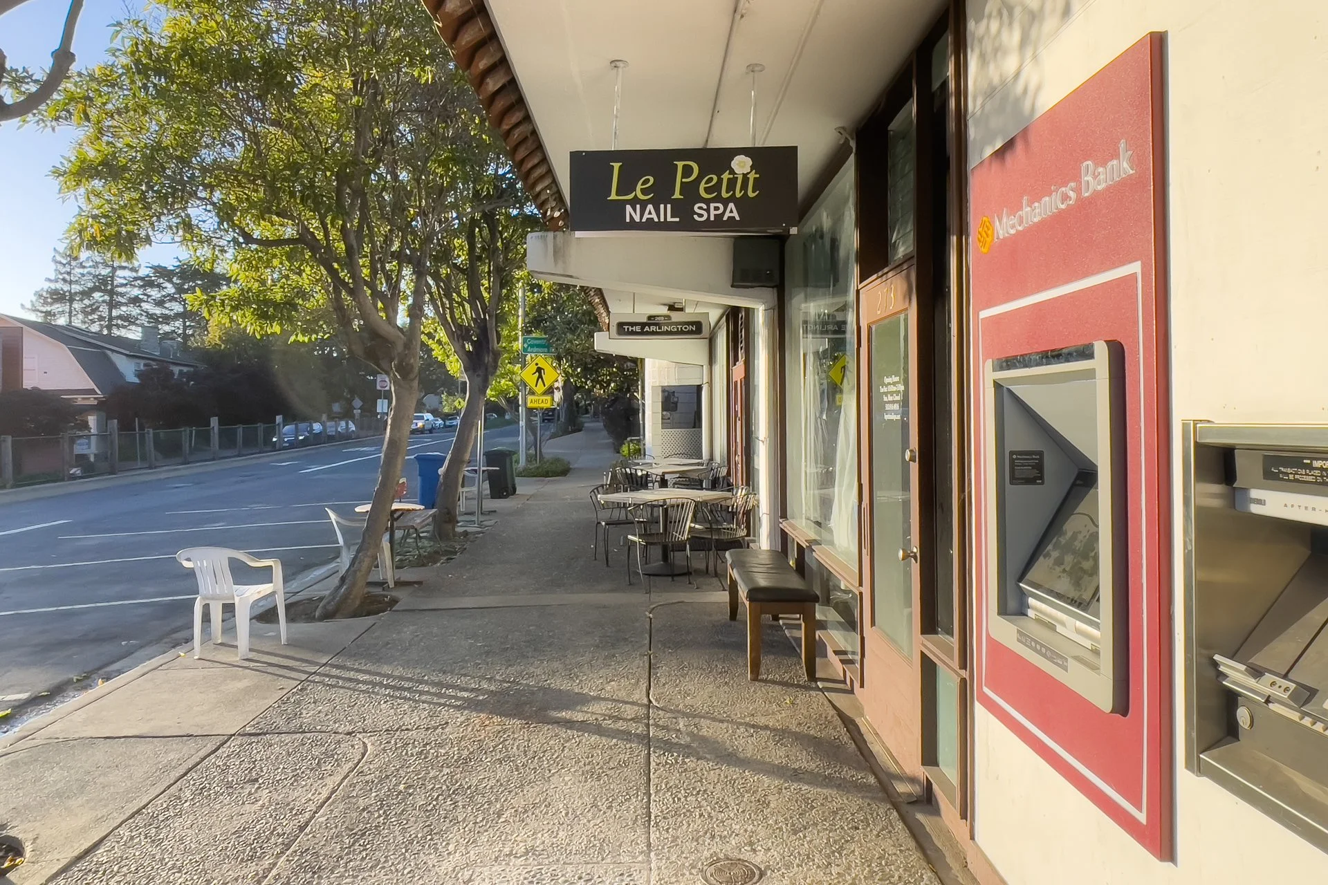 Sidewalk view outside of a nail spa with outdoor seating, ATM machine, ATM sign, trees, crosswalk sign, and parking lot.