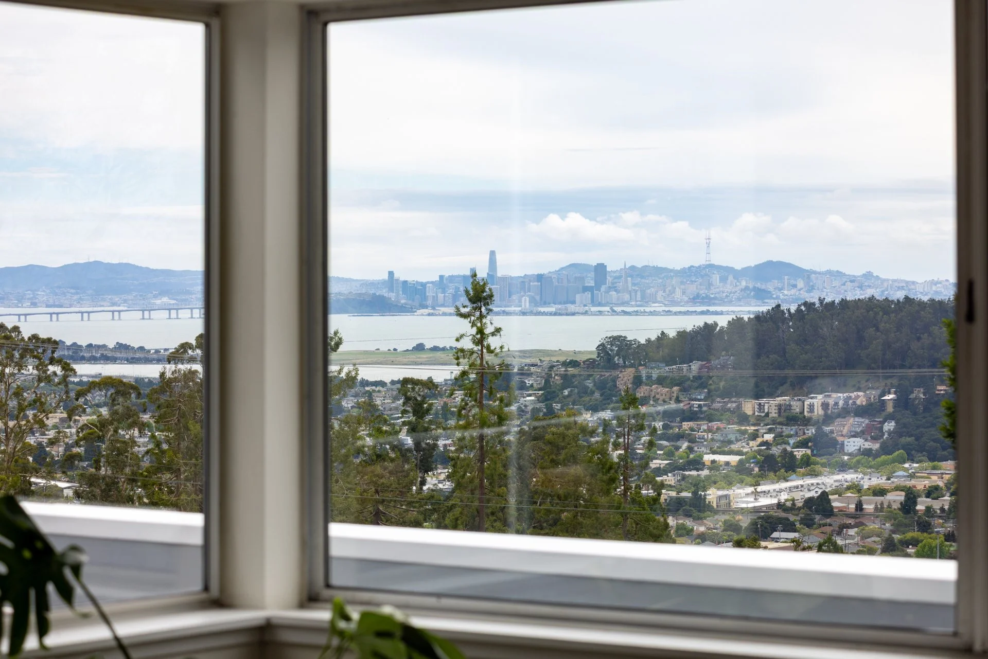 View of a city skyline with tall buildings, water, and hills in the distance, seen through an open window. There are some trees and residential areas in the foreground.