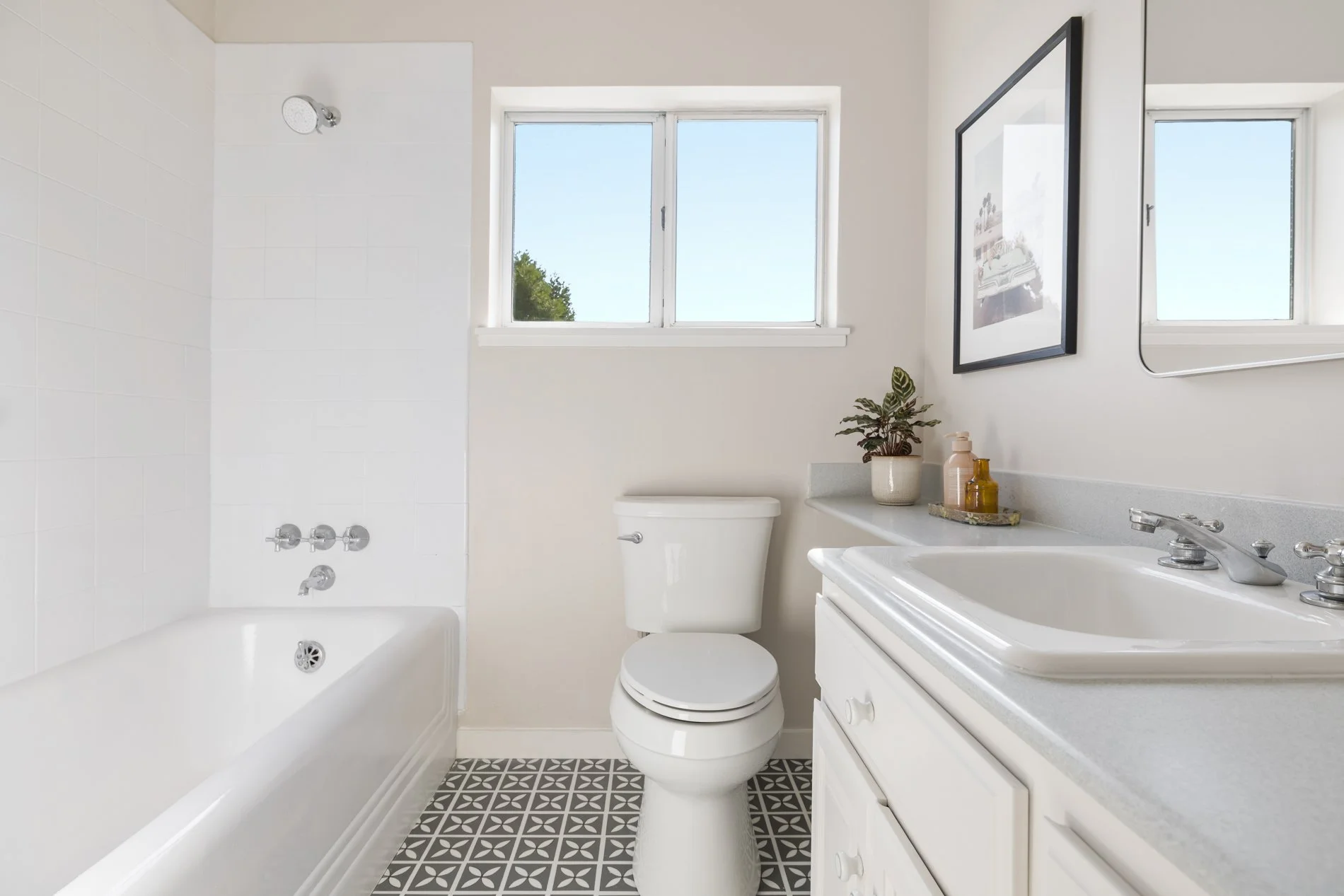 A clean, bright bathroom featuring a bathtub, a toilet, a white vanity with a sink, a mirror, a potted plant, and a window with a view of trees.