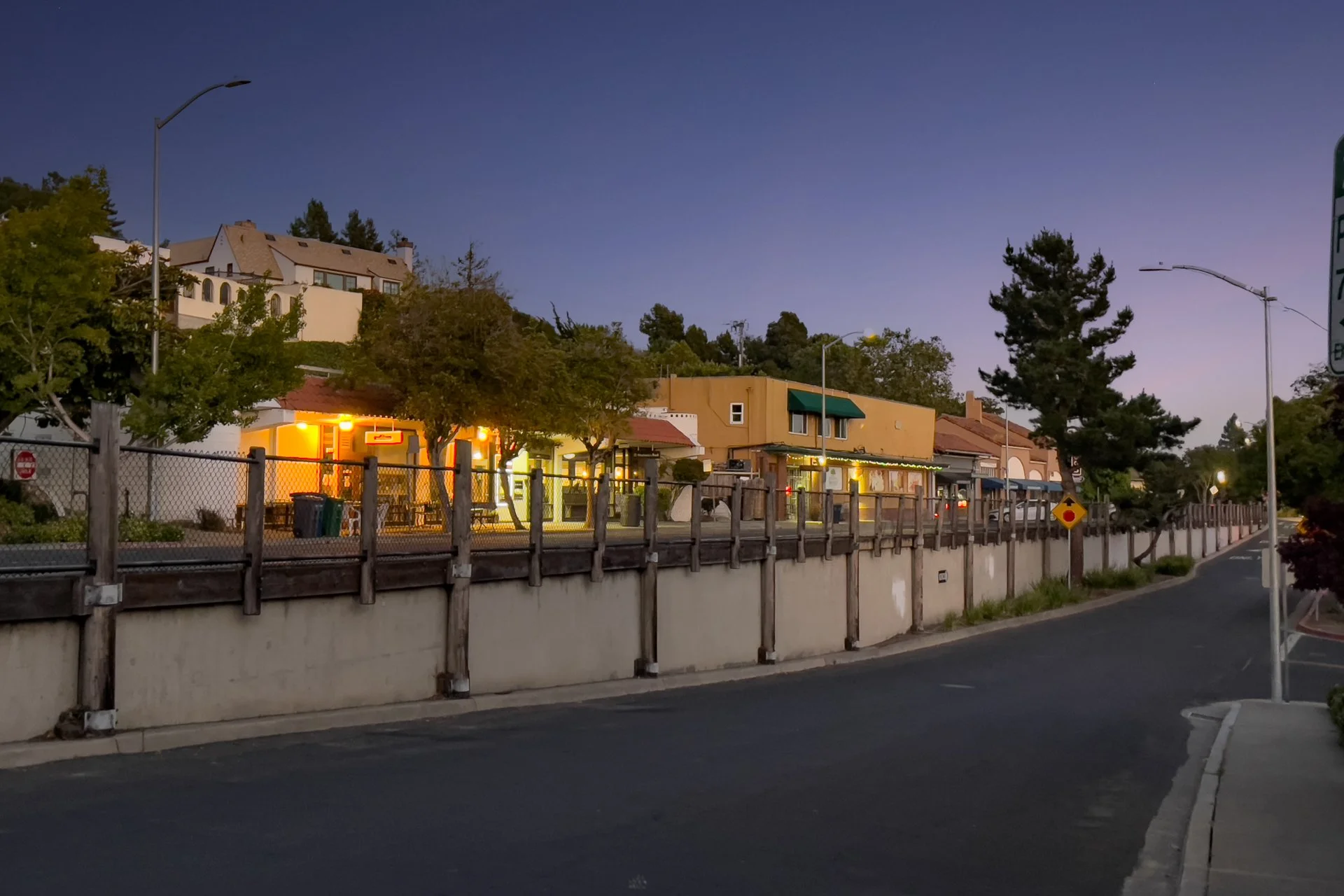 Dusk scene of a small street with store buildings and trees, some illuminated with yellow and white lights, against a darkening sky.