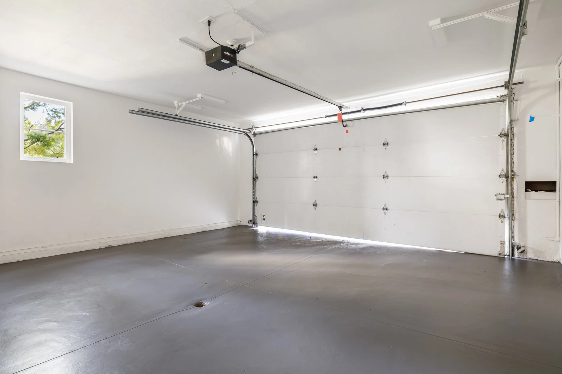 Empty residential garage with white walls, a closed white garage door, a small window showing trees outside, and a smooth gray concrete floor.