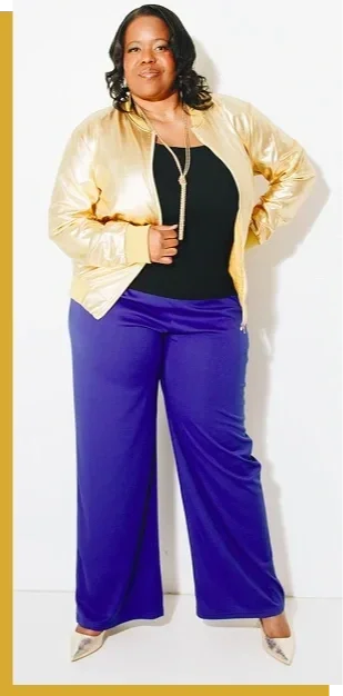 A woman standing against a plain white background, wearing a gold jacket, black top, purple pants, and beige heels, holding a long pearl necklace.