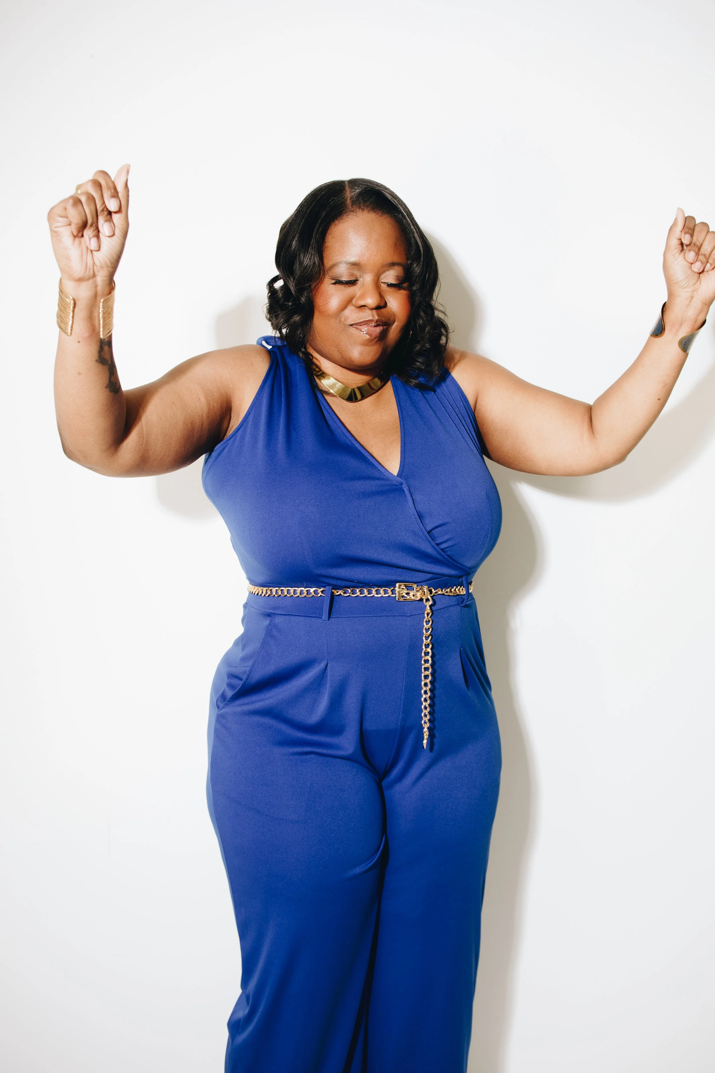 A woman with dark, shoulder-length wavy hair, wearing a sleeveless blue jumpsuit with a gold belt and jewelry, is dancing with her eyes closed and arms raised against a white background.