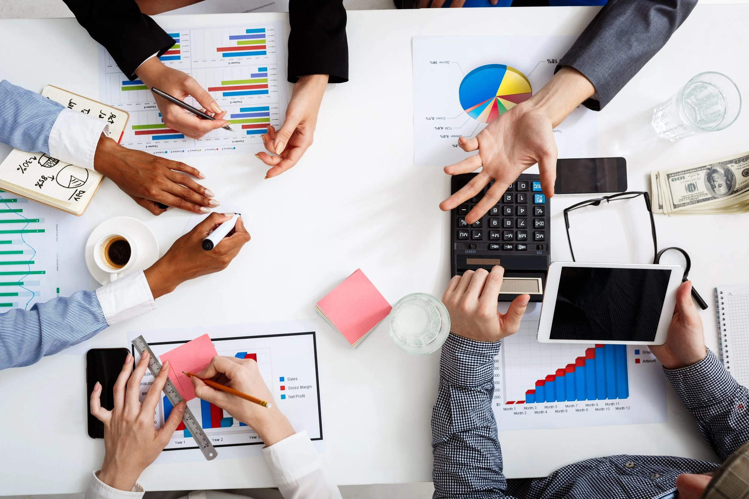 A top-down view of a business meeting with multiple people working on graphs, charts, and financial documents on a white table, including a calculator, glasses of water, a coffee cup, and various stationery items.