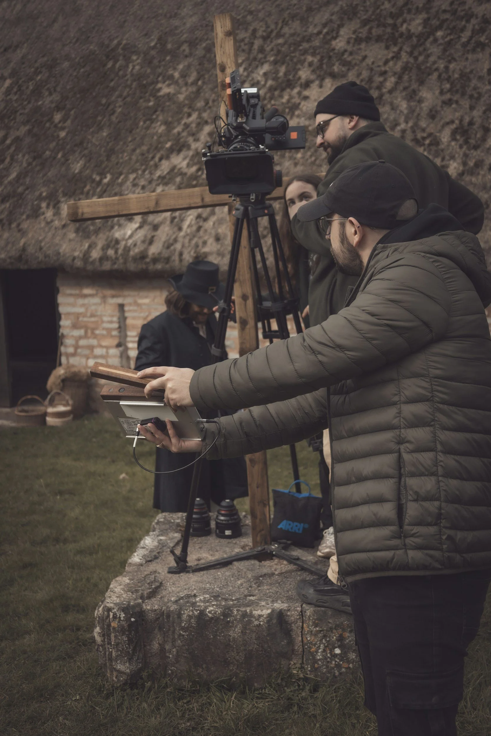 Group of people setting up a camera on a tripod outdoors during daytime, with a brick building and grassy area in background.