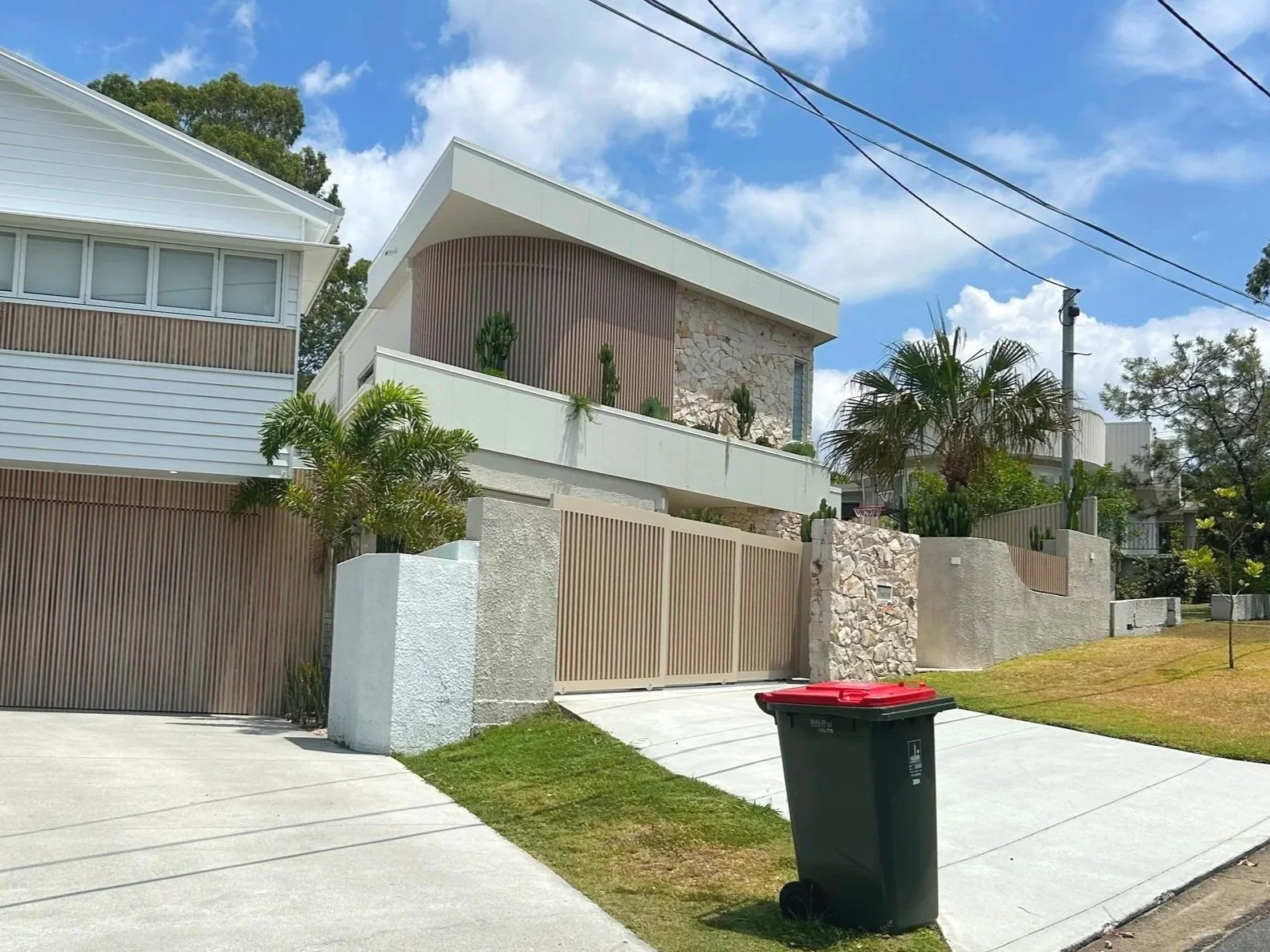 Modern house with white and beige exterior, surrounded by a concrete fence and gate, with tropical plants and palm trees in front, under a partly cloudy sky.