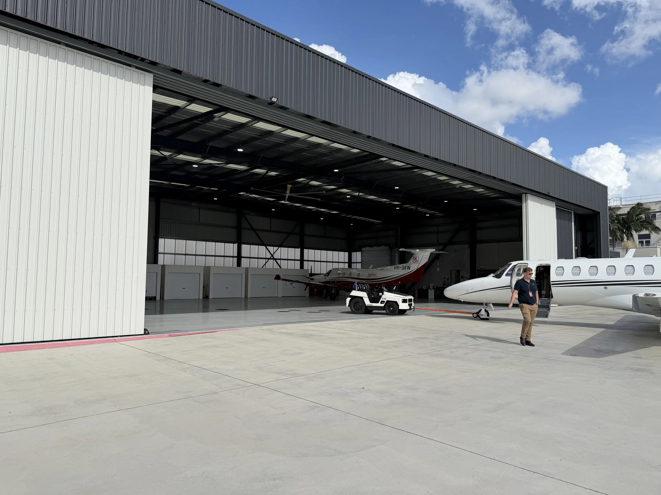 Inside a private hangar with a small aircraft parked, a ground crew member walking nearby, and open space with a view of blue sky and clouds.