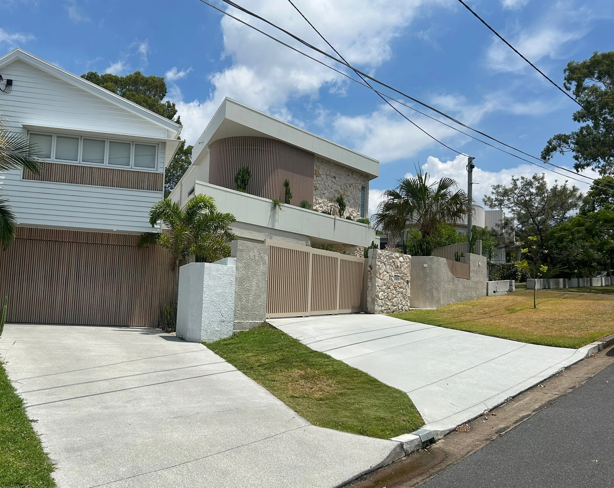 Modern house with white and beige exterior, surrounded by a concrete fence and gate, with tropical plants and palm trees in front, under a partly cloudy sky.