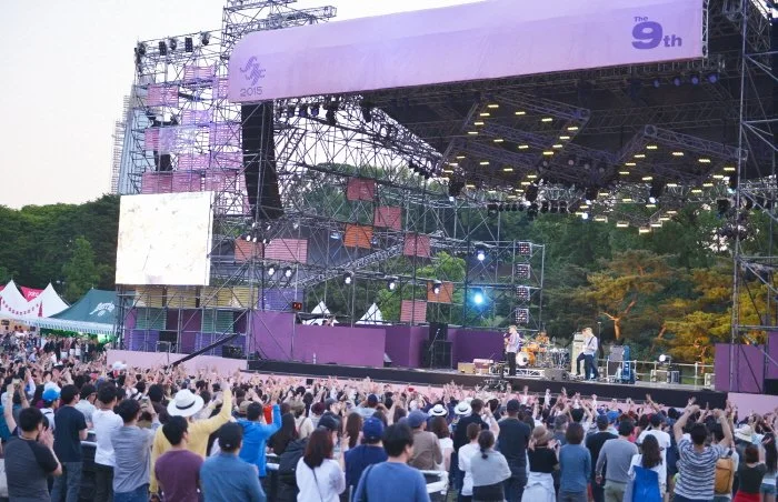 Outdoor concert stage with a large crowd watching live music performance in a park with trees, during daytime.