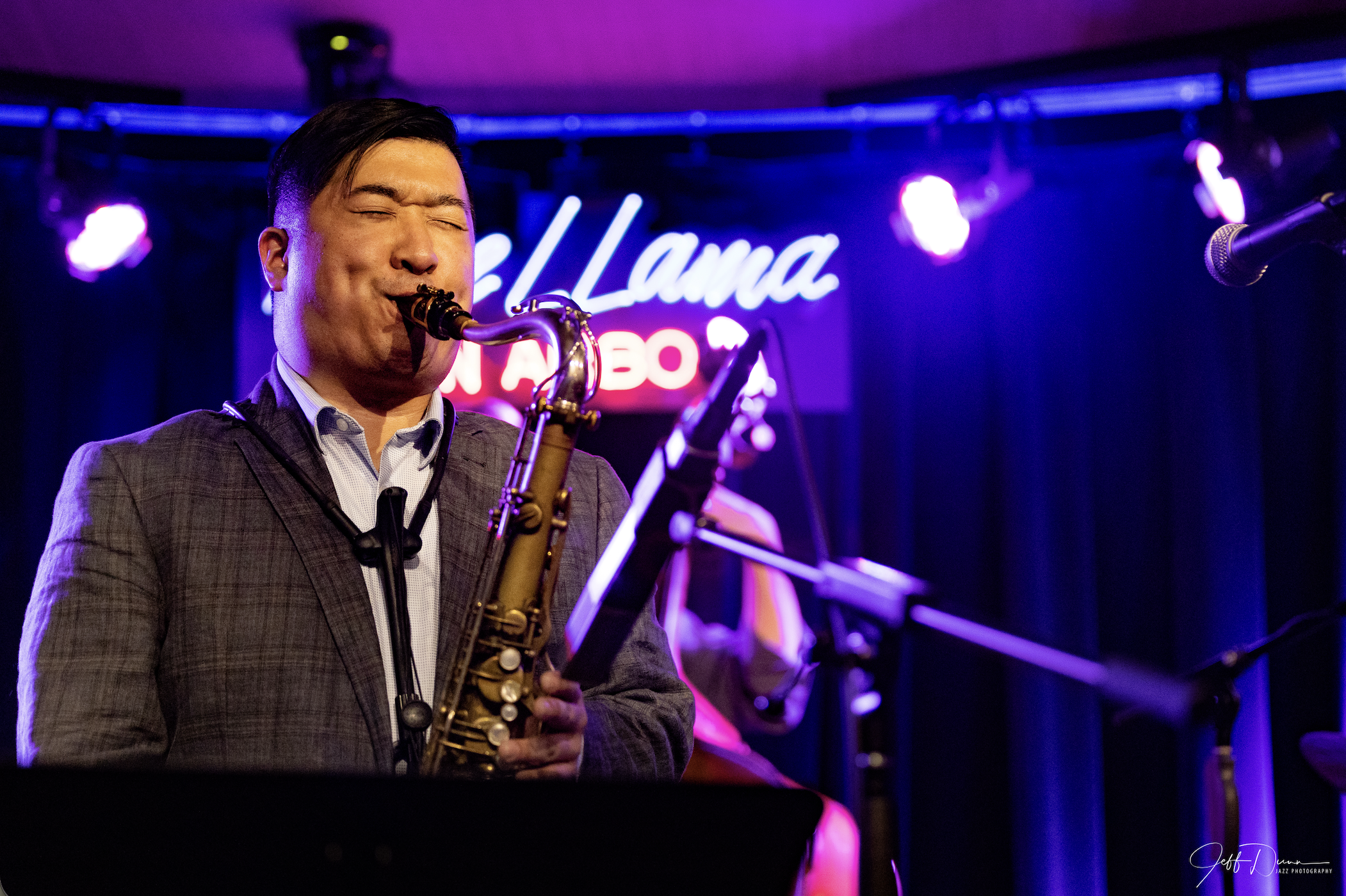 A man in a suit playing a saxophone on stage at a jazz club with a neon sign in the background reading 'Llama Jazz'.
