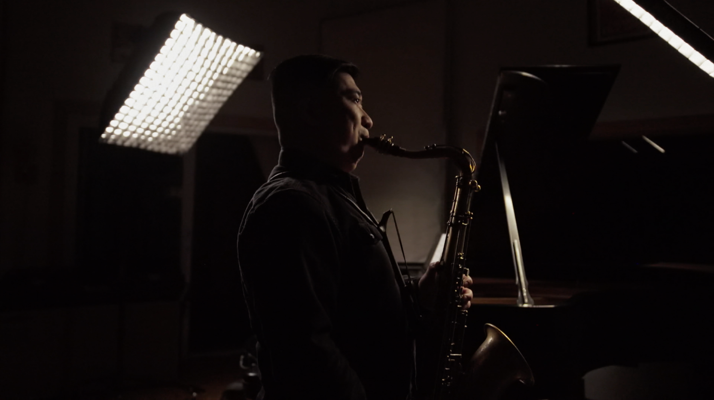 A man playing the saxophone in a dimly lit room with a piano and a large light panel above him.