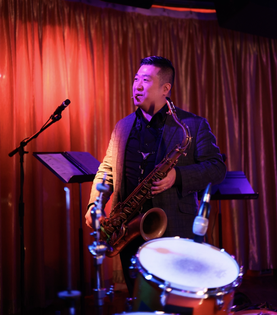 A musician holding a saxophone on stage, with a drum in the foreground and a microphone on a stand, against a backdrop of red curtains.