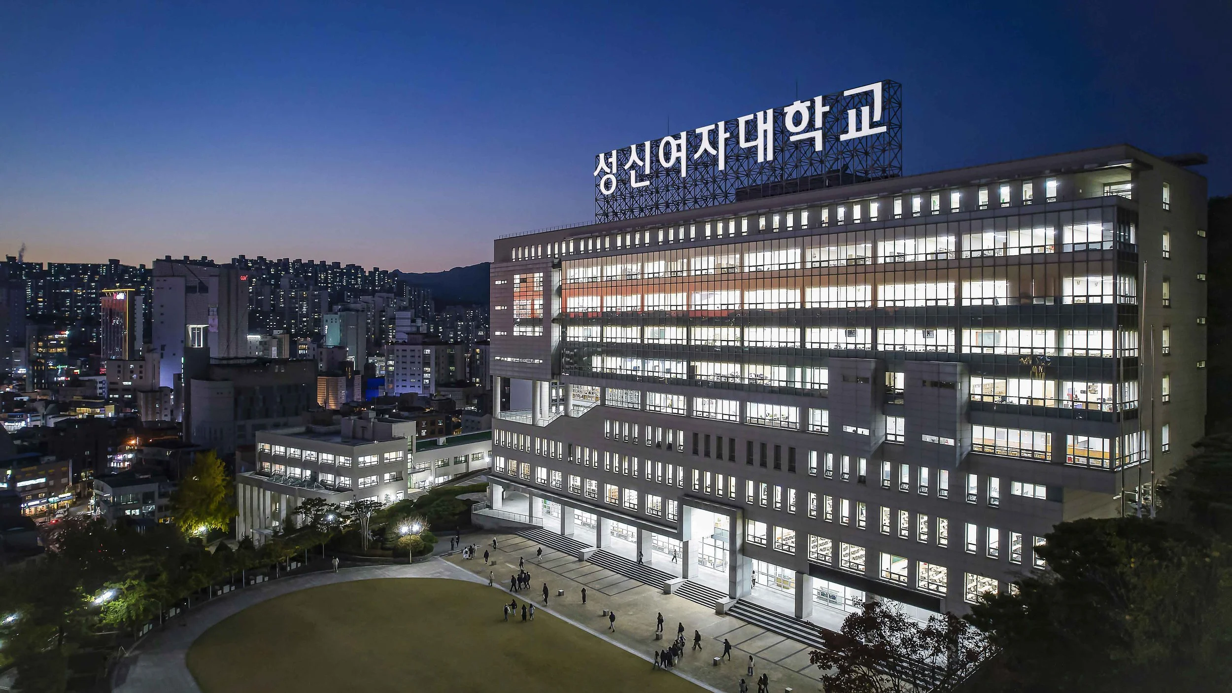 Nighttime cityscape featuring a large university building with illuminated windows, a sign in Korean on the roof, and a landscaped courtyard with people walking.