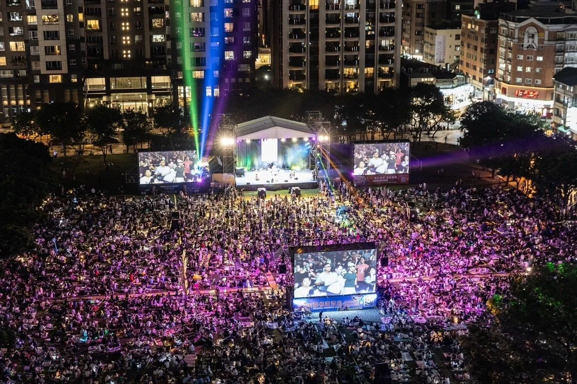An outdoor concert at night with a large audience, a stage with colorful lighting, and two big screens displaying the performers, surrounded by tall buildings.