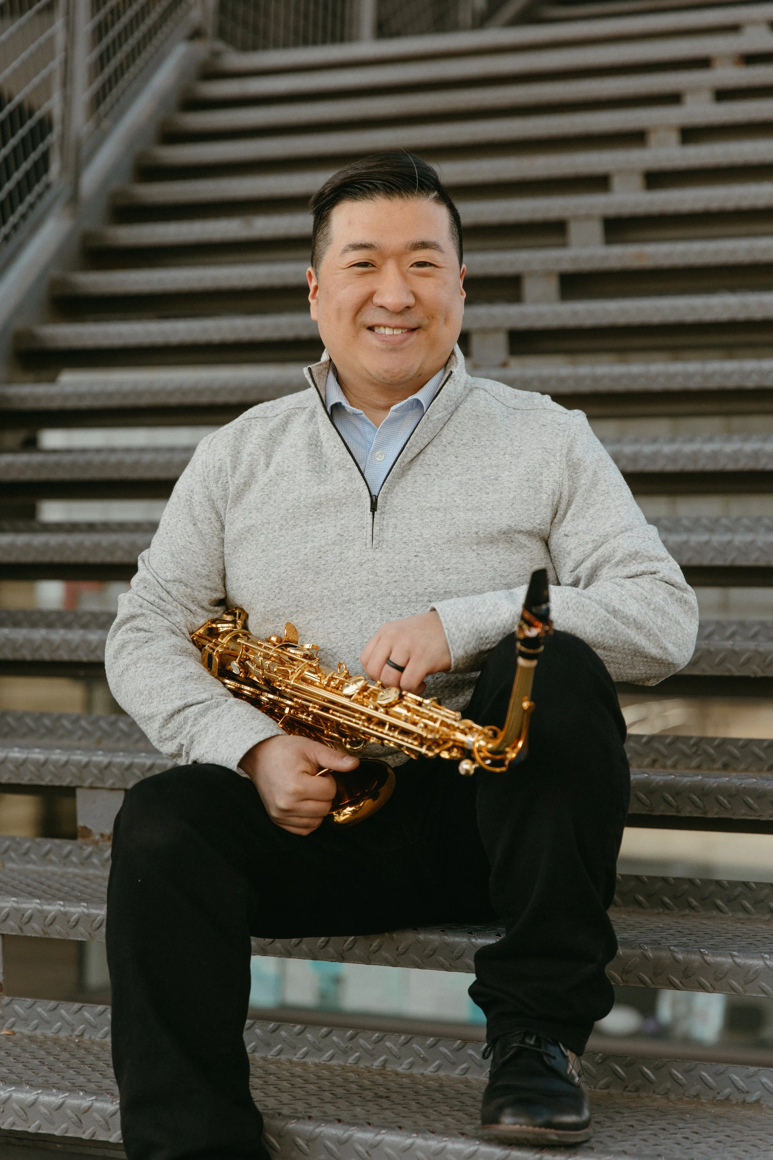 A man sitting on metal stairs holding a gold-colored saxophone, wearing a light gray zip-up jacket over a collared shirt, with short dark hair and smiling.