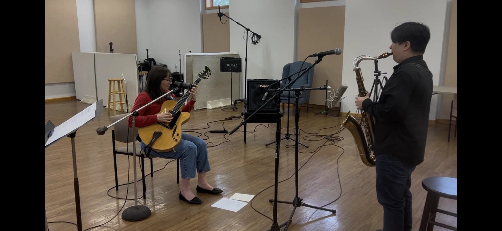 A woman playing an acoustic guitar and a man playing a saxophone in a music studio with microphones, music stands, and recording equipment around them.