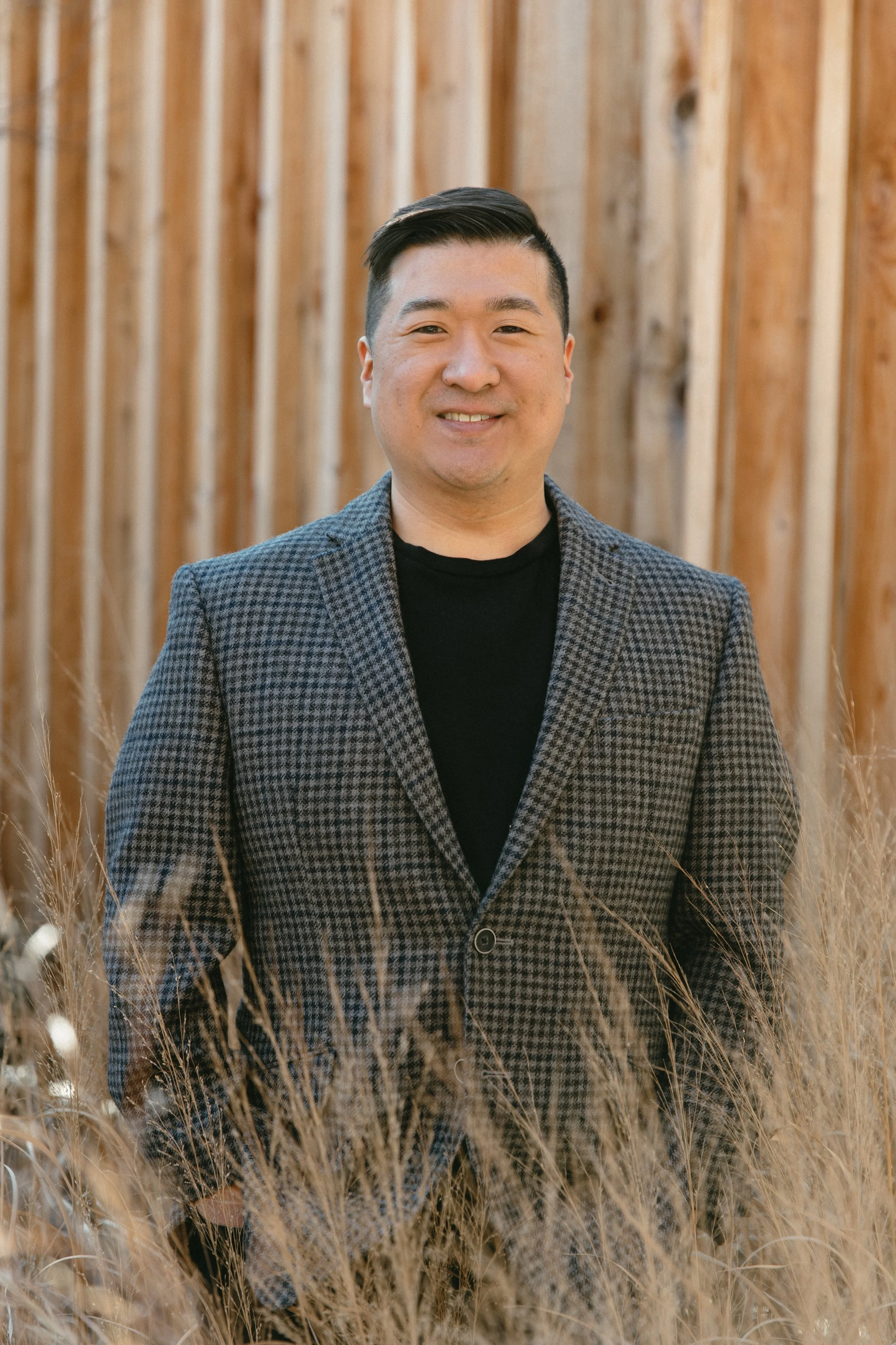A man in a checkered blazer and black t-shirt standing outdoors in front of a wooden fence, surrounded by tall, dry grass.
