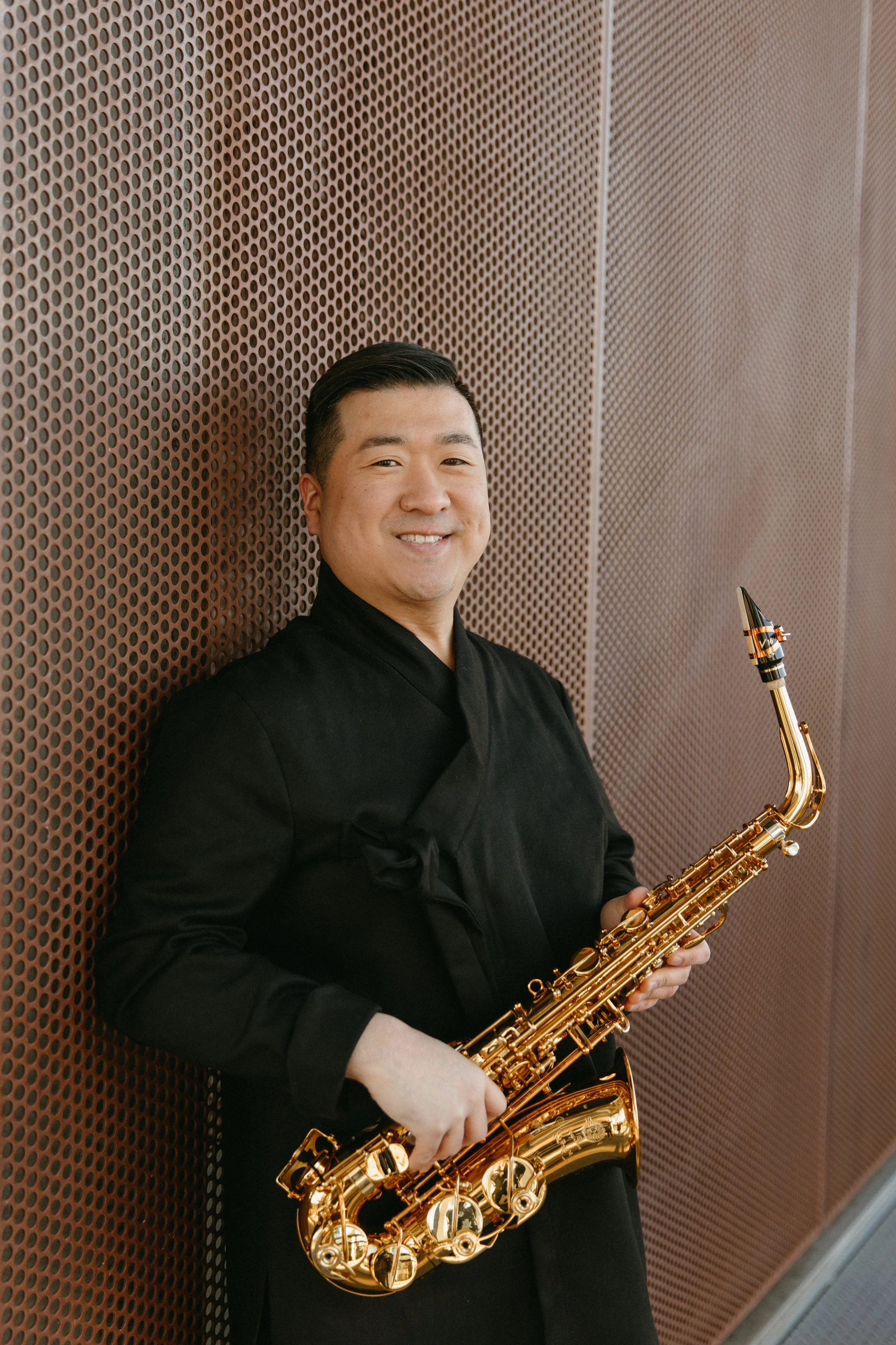 A man in black clothing holding a gold saxophone, standing against a brown perforated wall, smiling at the camera.