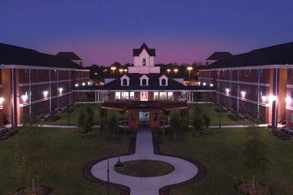 A large residential or hotel complex with a central courtyard, green lawn, and a pathway leading to a white building with a tower, under twilight sky.