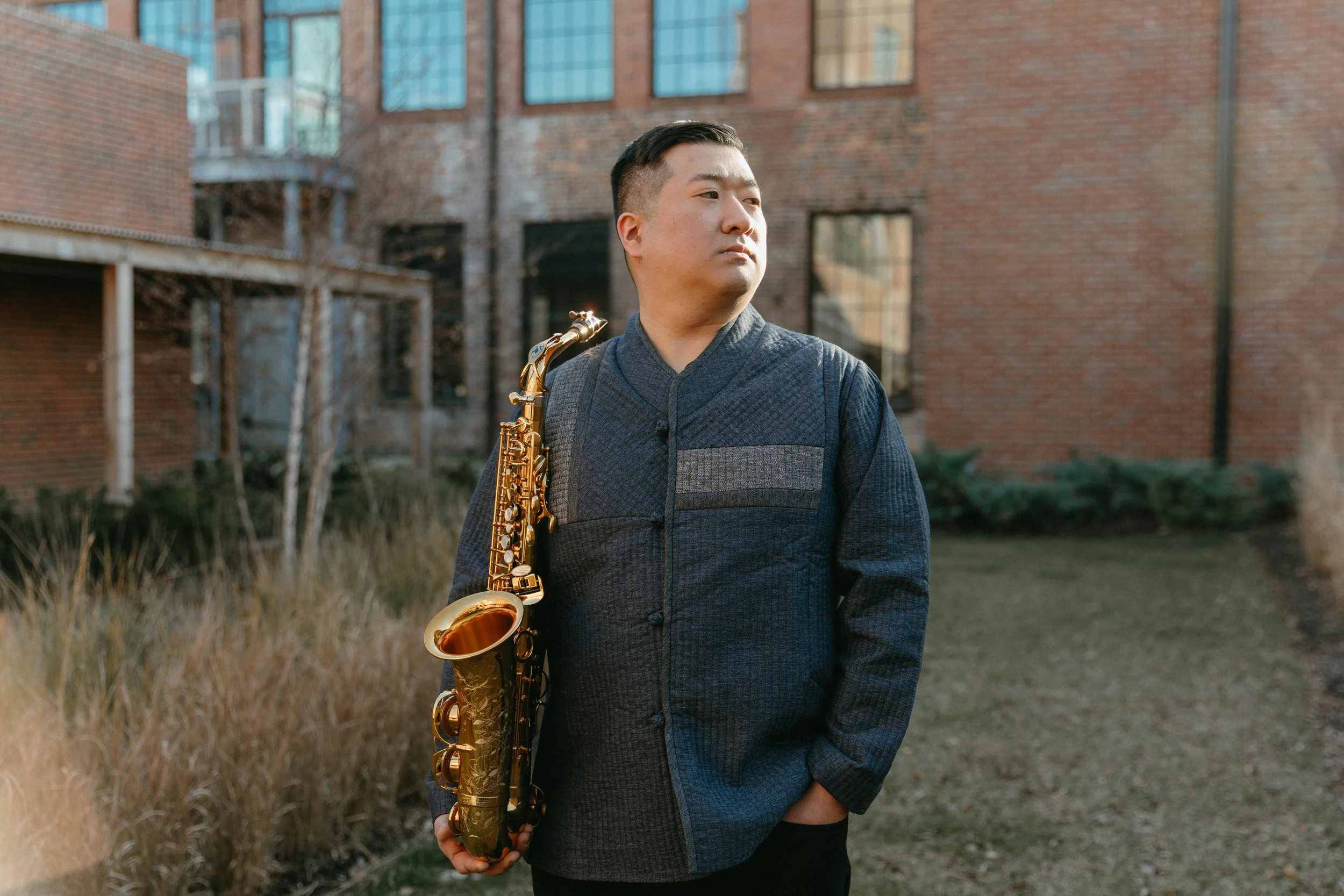 A man stands outdoors in front of brick buildings, holding a gold-colored saxophone over his shoulder and looking to his left.