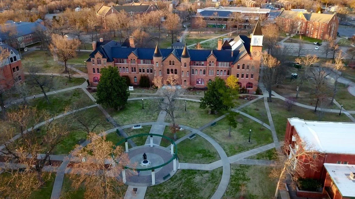 An aerial view of a historic brick building with pointed towers, surrounded by a park with walkways, benches, and trees, in a college campus setting.