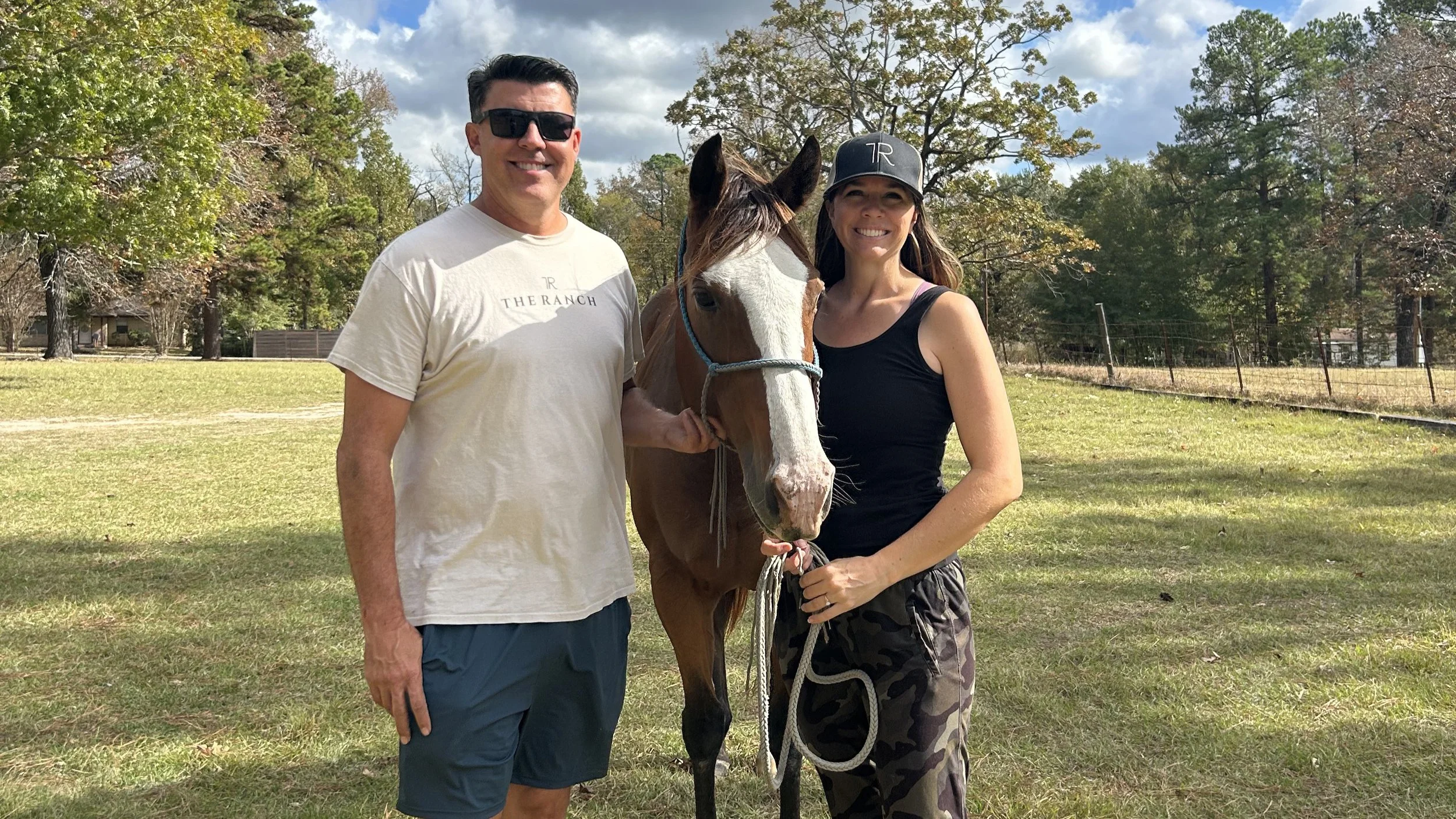 A man and a woman standing next to each other outdoors with a horse between them, smiling at the camera, in a grassy area with trees in the background.