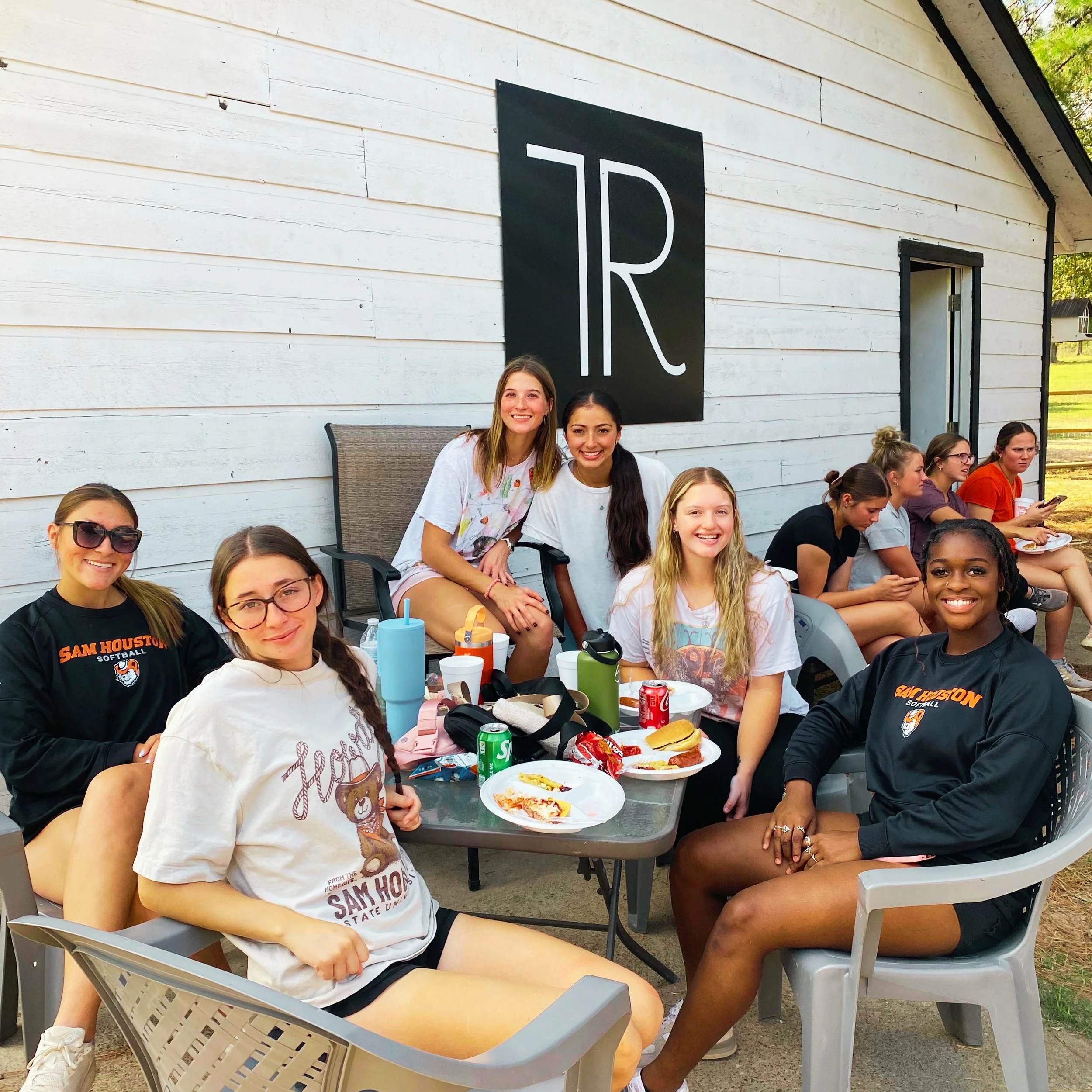 A group of diverse young women sitting outside at a table, smiling, with food and drinks, near a white building with black accents and a large black sign with white lettering.