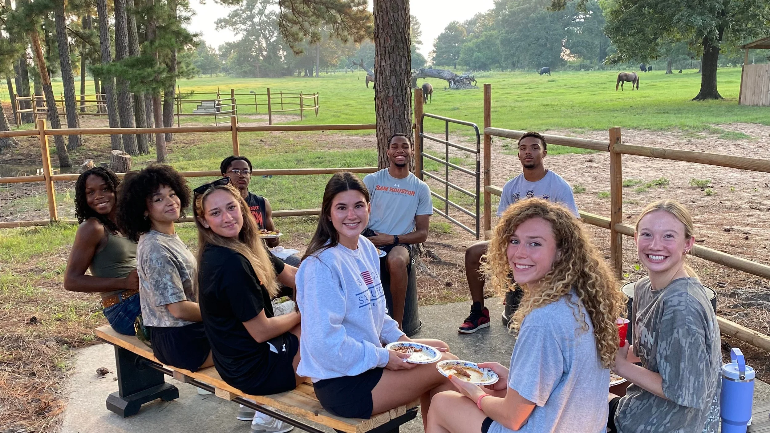 A group of nine young people sitting at a picnic table outdoors in a wooded area, with a grassy field and trees in the background, and several horses grazing in the distance.